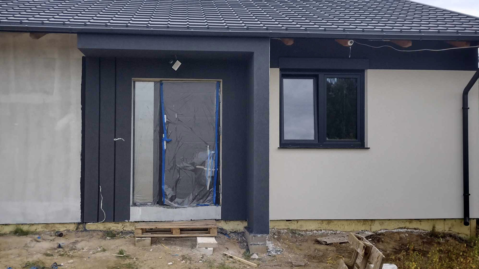 Fragment elewacji domu w trakcie prac wykończeniowych: szara stolarka okienna i drzwiowa, częściowo otynkowana ściana w kolorze złamanej bieli, zabezpieczone drzwi.