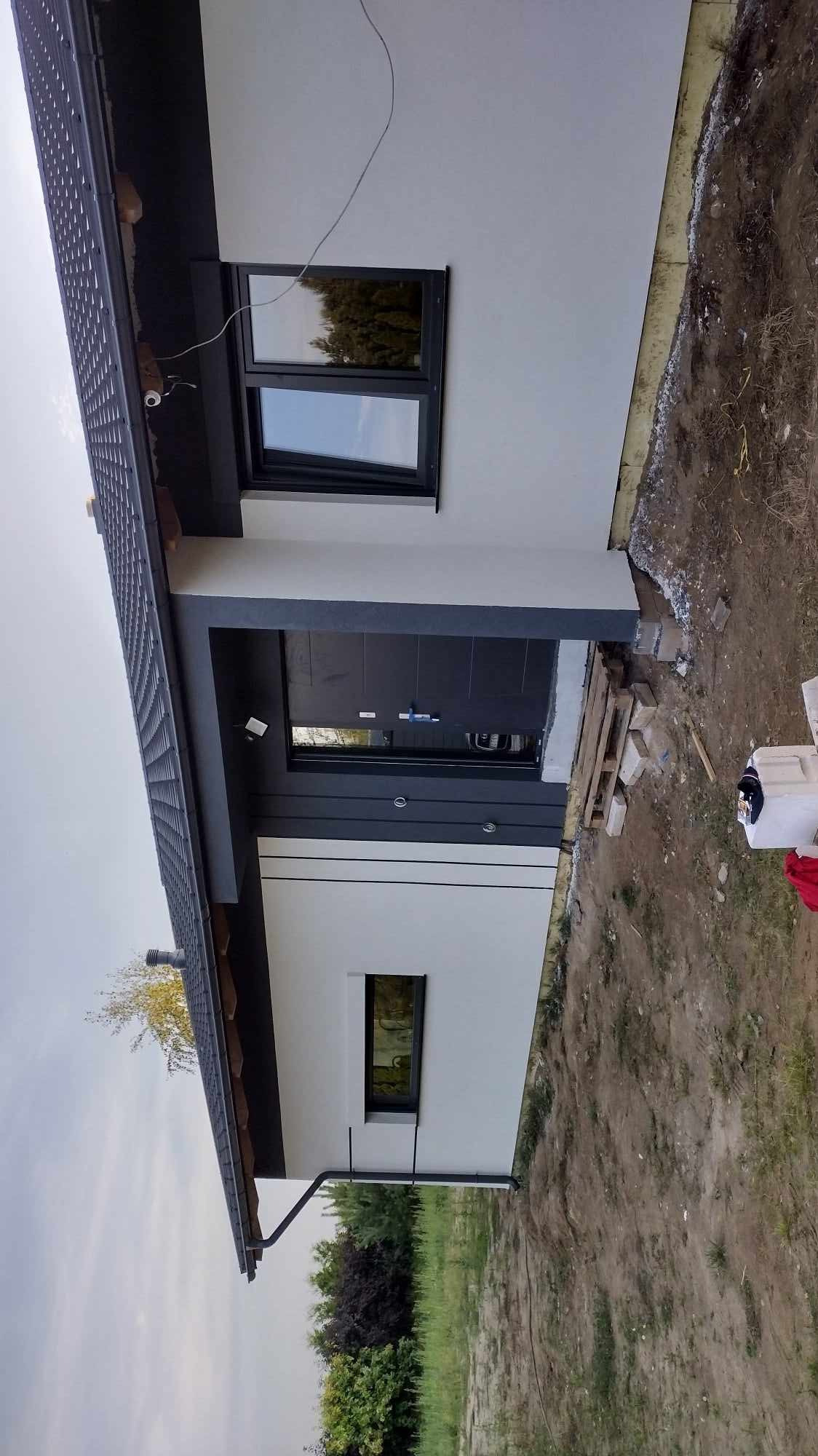 Nowoczesny dom jednorodzinny z szarą stolarką okienną i drzwiową, białą elewacją oraz fragmentem ocieplenia widocznym przy gruncie. Widoczna boczna elewacja budynku.