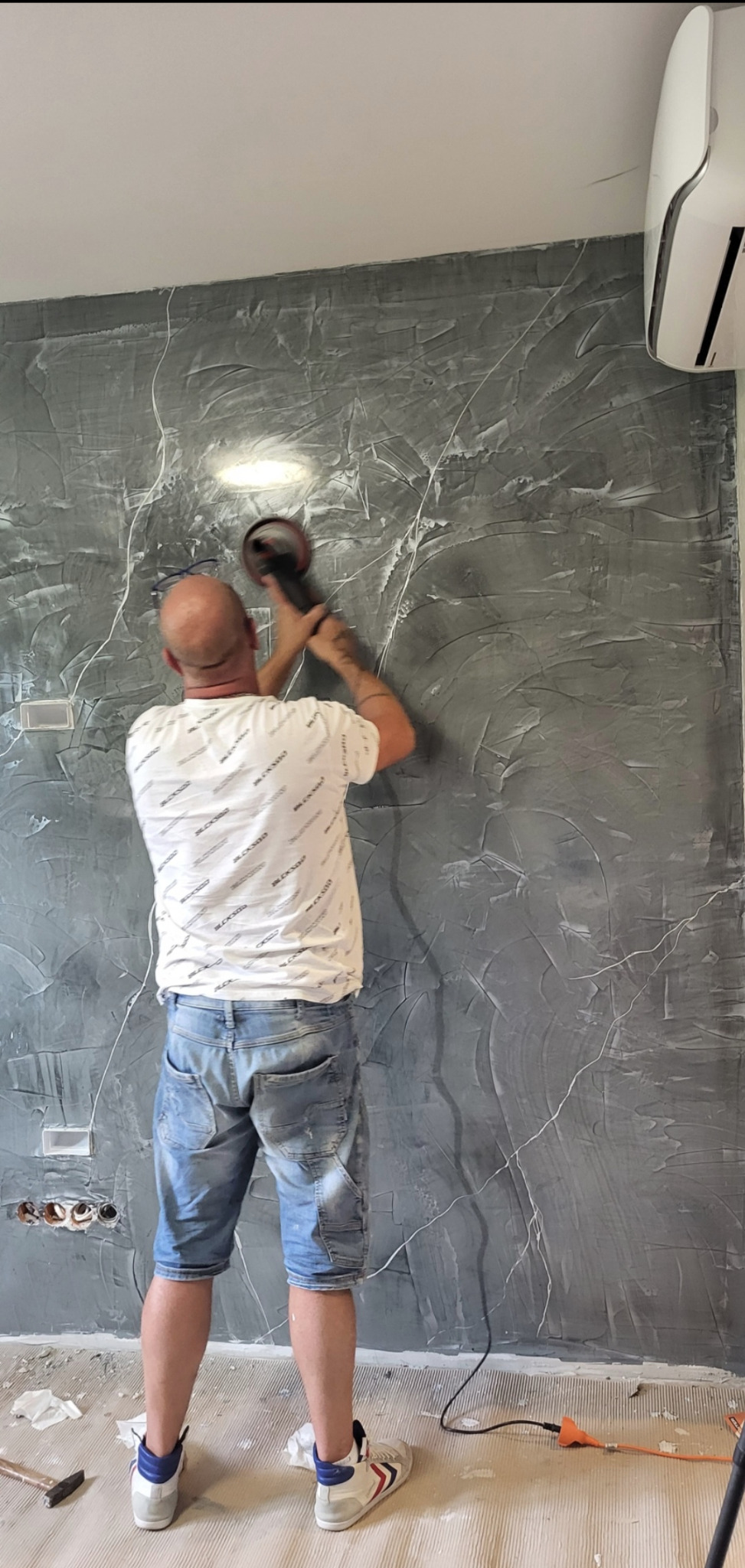 Pracownik szlifuje ścianę z dekoracyjną szarą strukturą przypominającą marmur, używając szlifierki. Widoczne gniazdka elektryczne i klimatyzator.