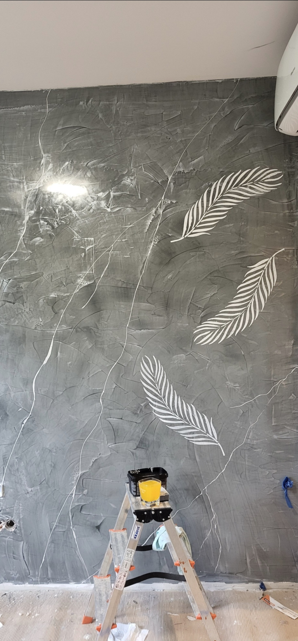 Szara ściana z efektem betonu i białymi, malowanymi piórami. Na drabinie stoi puszka z farbą i szklanka z pomarańczowym płynem. Widoczne gniazdka elektryczne.