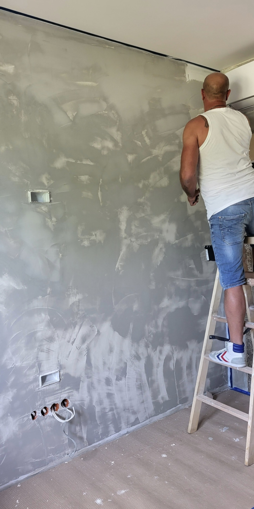Pracownik na drabinie aplikuje dekoracyjną, szarą farbę na ścianę w pomieszczeniu. Widoczne puszki elektryczne i zabezpieczona taśmą listwa sufitowa.