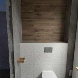 Wykończona toaleta z białymi, fakturowanymi i drewnopodobnymi płytkami. Widoczny drewniany uchwyt na papier toaletowy i fragment surowego muru przy wejściu.