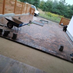 Tadeusz Gaca - Układanie kostki brukowej na posesji z drewnianym ogrodzeniem. Widoczny wózek, narzędzia i częściowo ułożona nawierzchnia. W tle biały bus i zieleń.