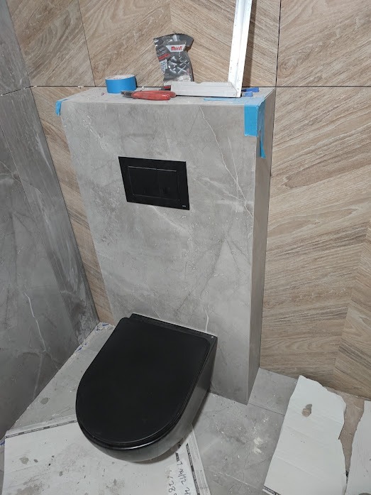 Podwieszana toaleta w zabudowie z płytek imitujących marmur, czarny przycisk spłukiwania, narzędzia glazurnicze na górze, płytki na ścianach w dwóch odcieniach.