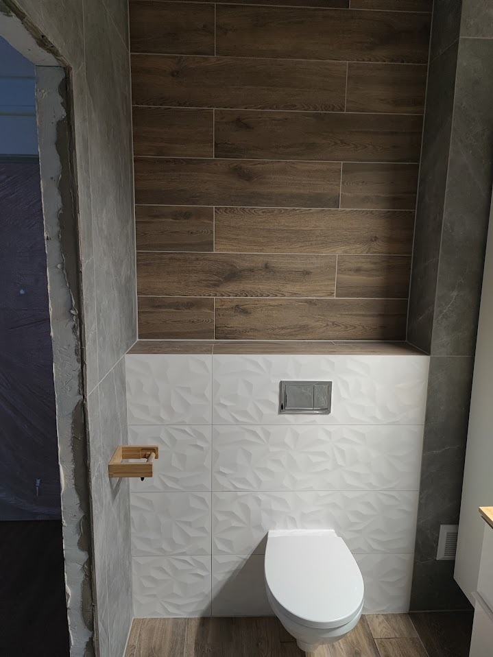 Wykończona toaleta z białymi, fakturowanymi i drewnopodobnymi płytkami. Widoczny drewniany uchwyt na papier toaletowy i fragment surowego muru przy wejściu.