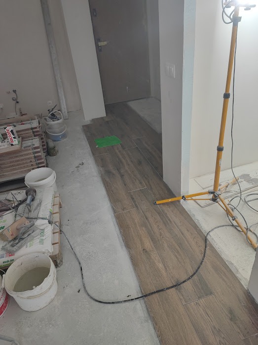 Układanie płytek imitujących drewno na podłodze w trakcie remontu. Widoczne narzędzia, materiały budowlane i lampa oświetleniowa. Częściowo ułożona podłoga.