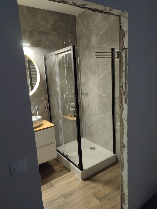 Nowoczesna kabina prysznicowa z czarnymi ramami, umywalka z szafką i lustro z podświetleniem w wykończonej łazience z szarą glazurą i panelami podłogowymi.