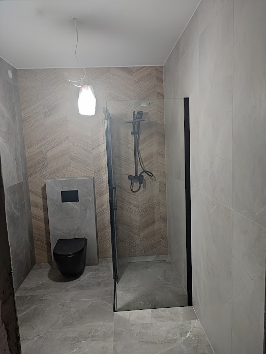 Wykończona łazienka z kabiną typu walk-in z czarnymi profilami, wiszącą miską WC w kolorze czarnym i płytkami imitującymi drewno na ścianie.