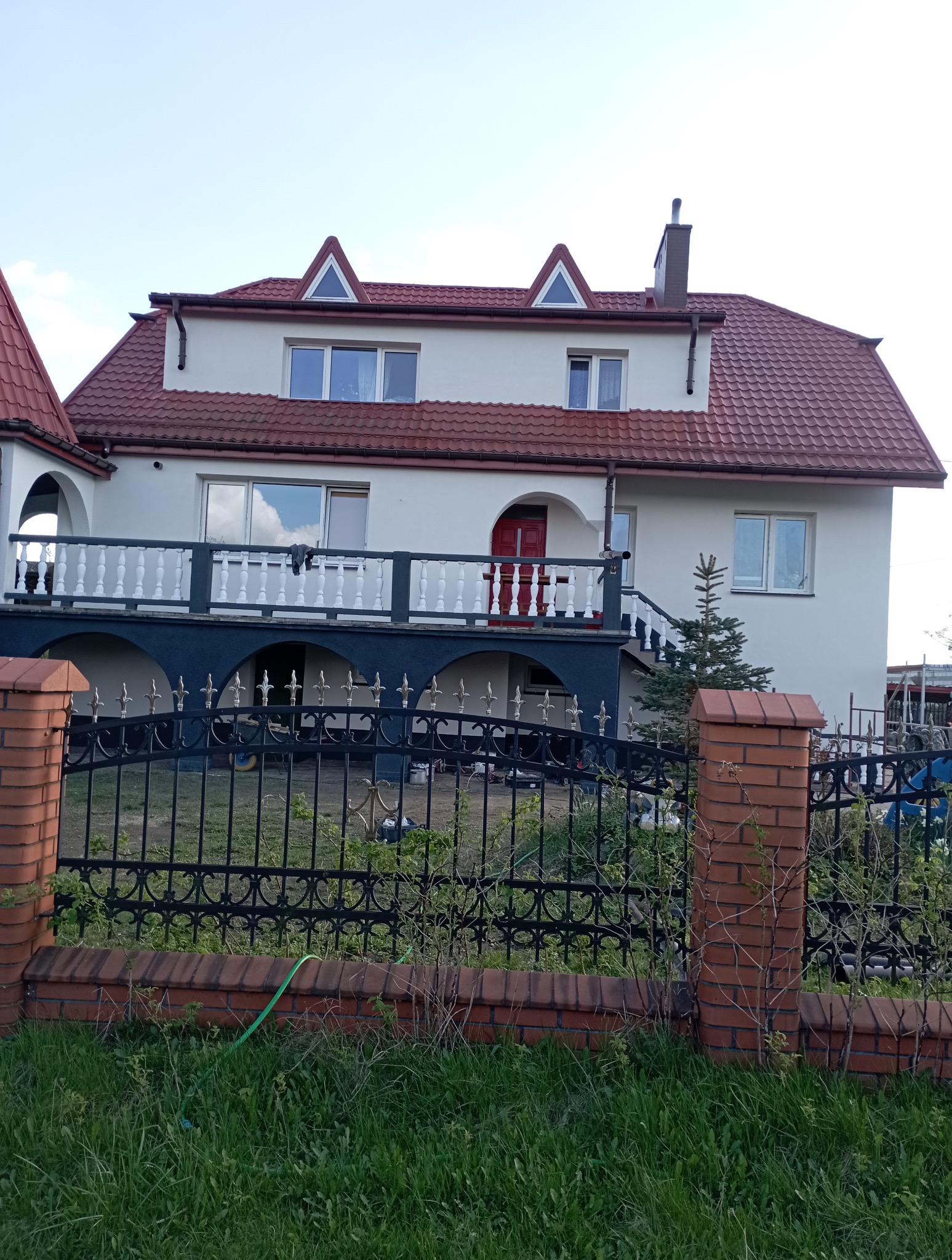 Dwupiętrowy dom z czerwonym dachem i balkonem, widziany zza ogrodzenia. Czerwone ceglane słupki ogrodzenia, bujna trawa. Dom pomalowany na jasny kolor z czerwonymi akcentami.