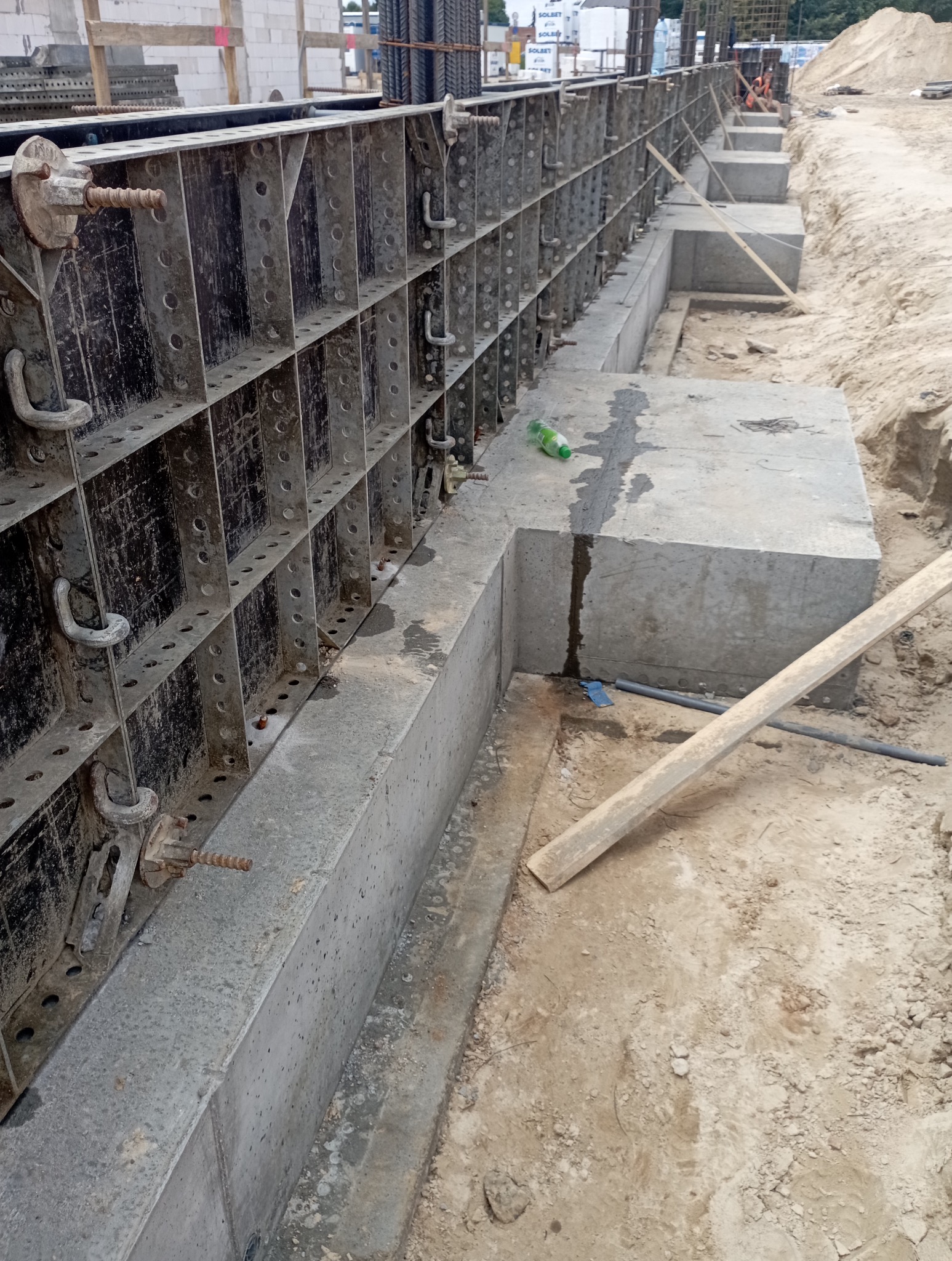 Szczegółowe ujęcie fundamentów z szalunkiem systemowym na placu budowy. Widoczne zbrojenie i betonowe elementy. Na pierwszym planie butelka po napoju.