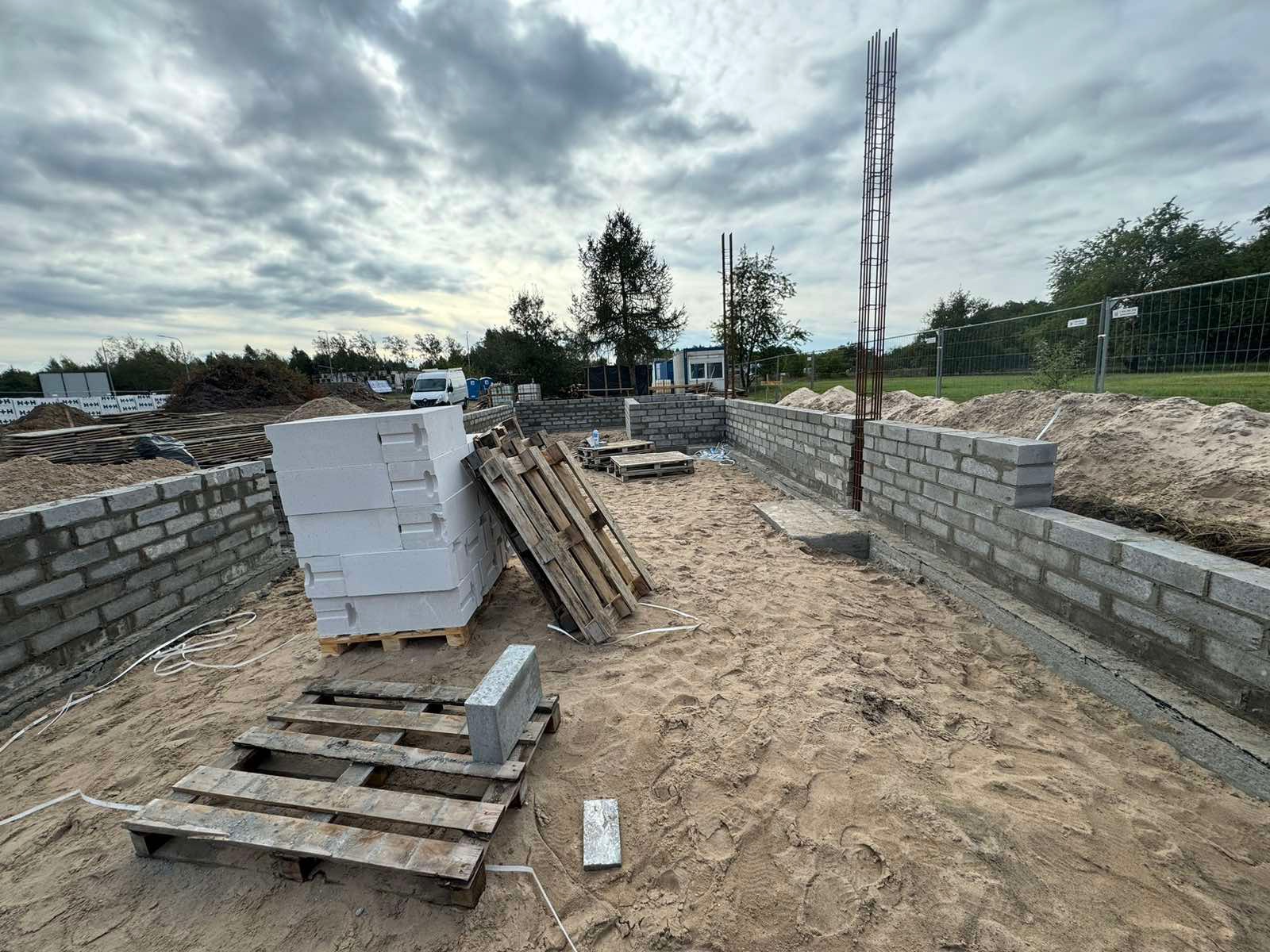 Wznoszenie murów fundamentowych z bloczków betonowych. Widoczne zbrojenia pionowe, palety z materiałami budowlanymi i piasek. Etap budowy domu przy pochmurnym niebie.