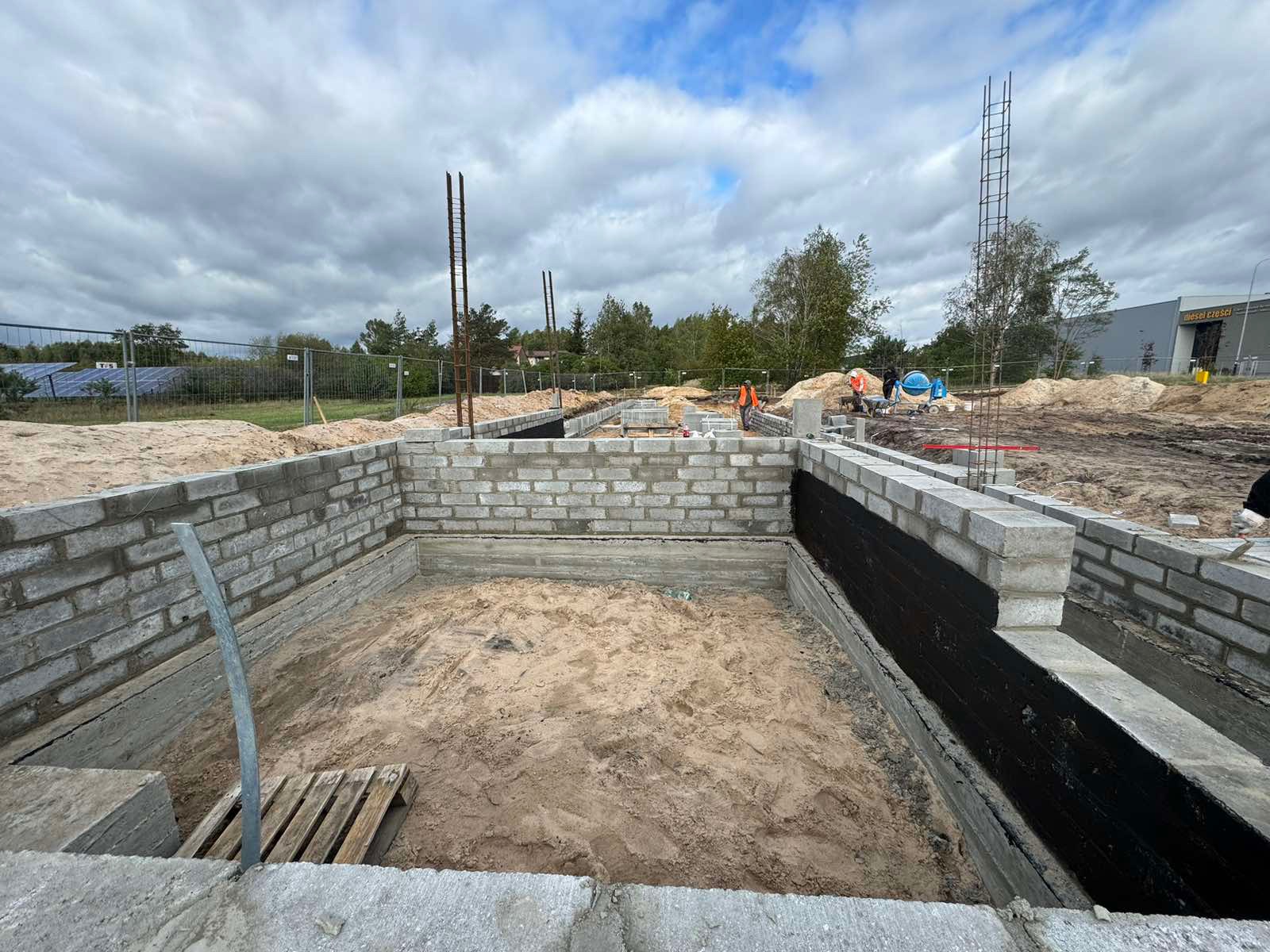 Fundamenty z bloczków betonowych, izolowane czarną folią, zbrojenie pionowe, piasek wewnątrz, widok na plac budowy i niebo z chmurami.