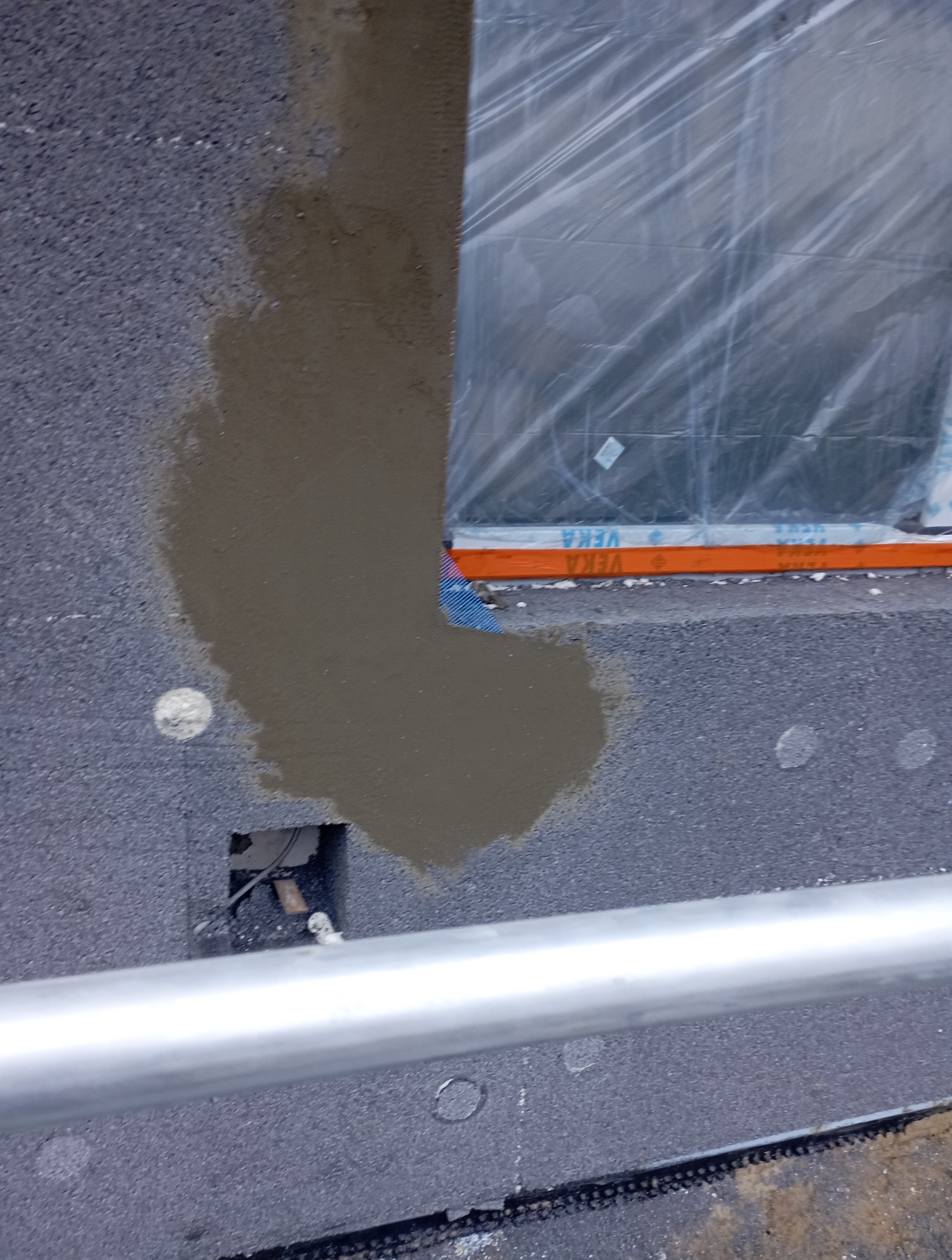 Aplikacja zaprawy na szary styropian przy oknie zabezpieczonym folią, widoczny fragment barierki i otwór instalacyjny w ociepleniu.