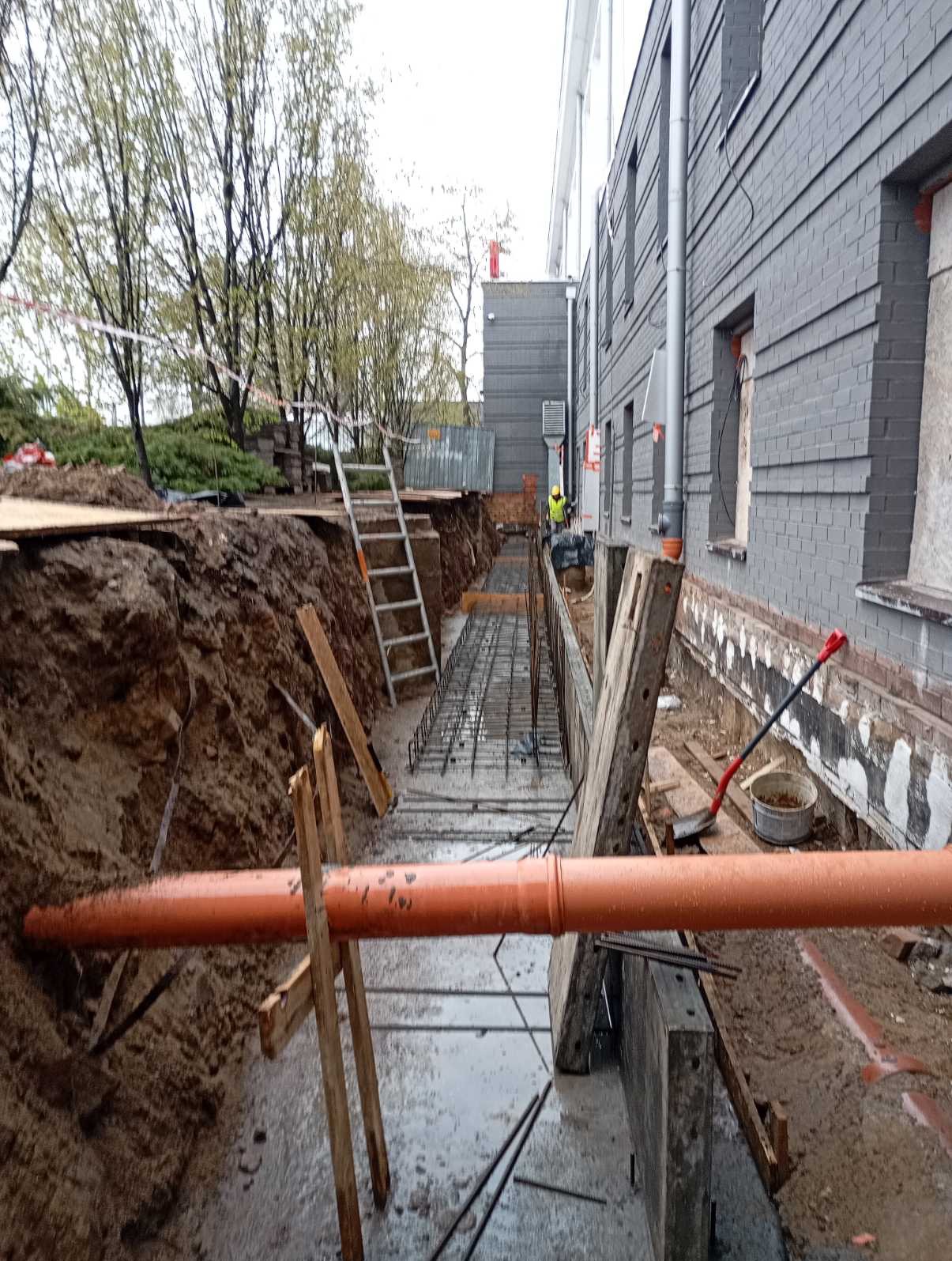 Wykop pod fundamenty budynku z ułożonym zbrojeniem i rurą kanalizacyjną. Widoczne szalunki i drabina. Budynek z szarej cegły w tle.