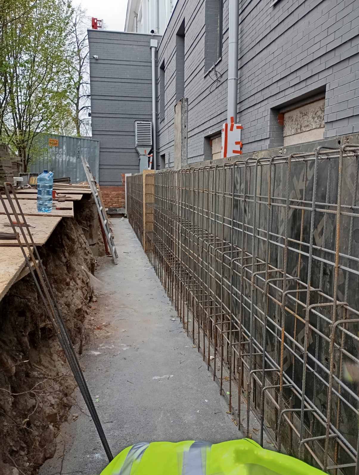 Zbrojenie fundamentów budynku z szarej cegły, widoczne z wykopu. Na pierwszym planie fragment żółtej kamizelki odblaskowej, w tle drabina i butelka wody.