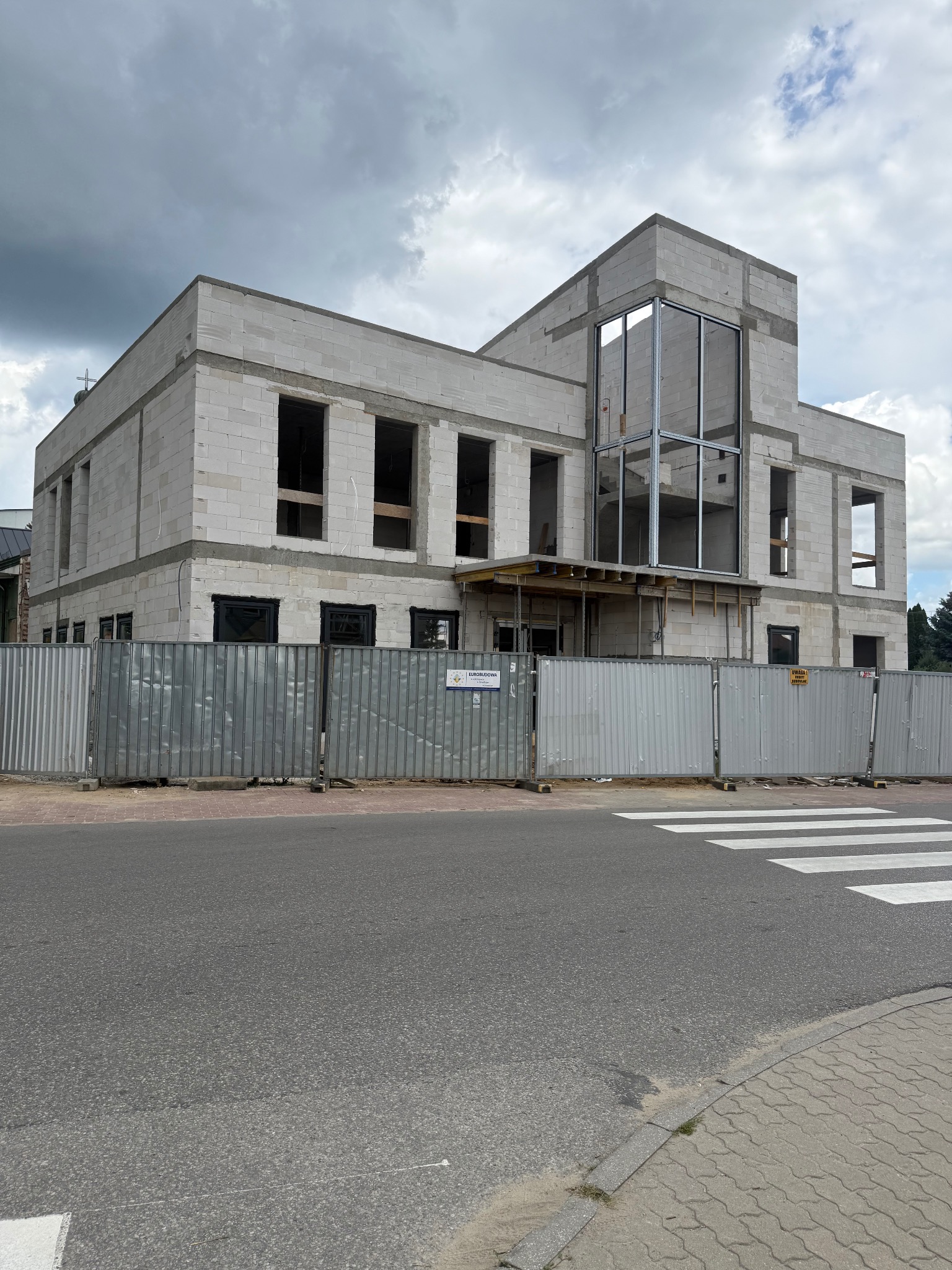 Budowa dwukondygnacyjnego budynku z białego betonu komórkowego, widok z zewnątrz, ogrodzenie budowy, stan surowy otwarty, okna bez szyb.