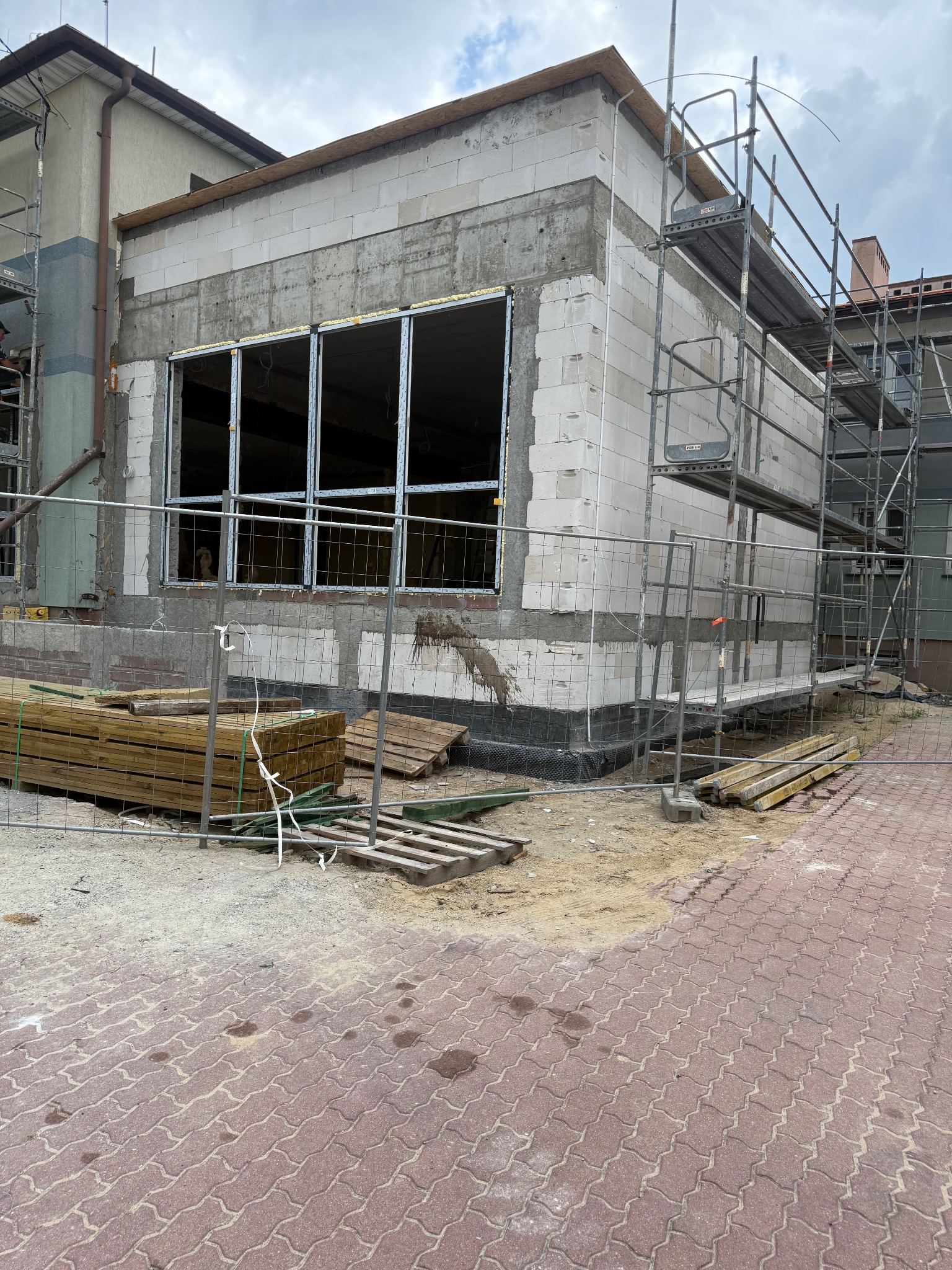 Budowa domu: ściany z białych bloczków, okna bez szyb, rusztowania i ogrodzenie placu budowy. Widoczne materiały budowlane i fragment chodnika z kostki.