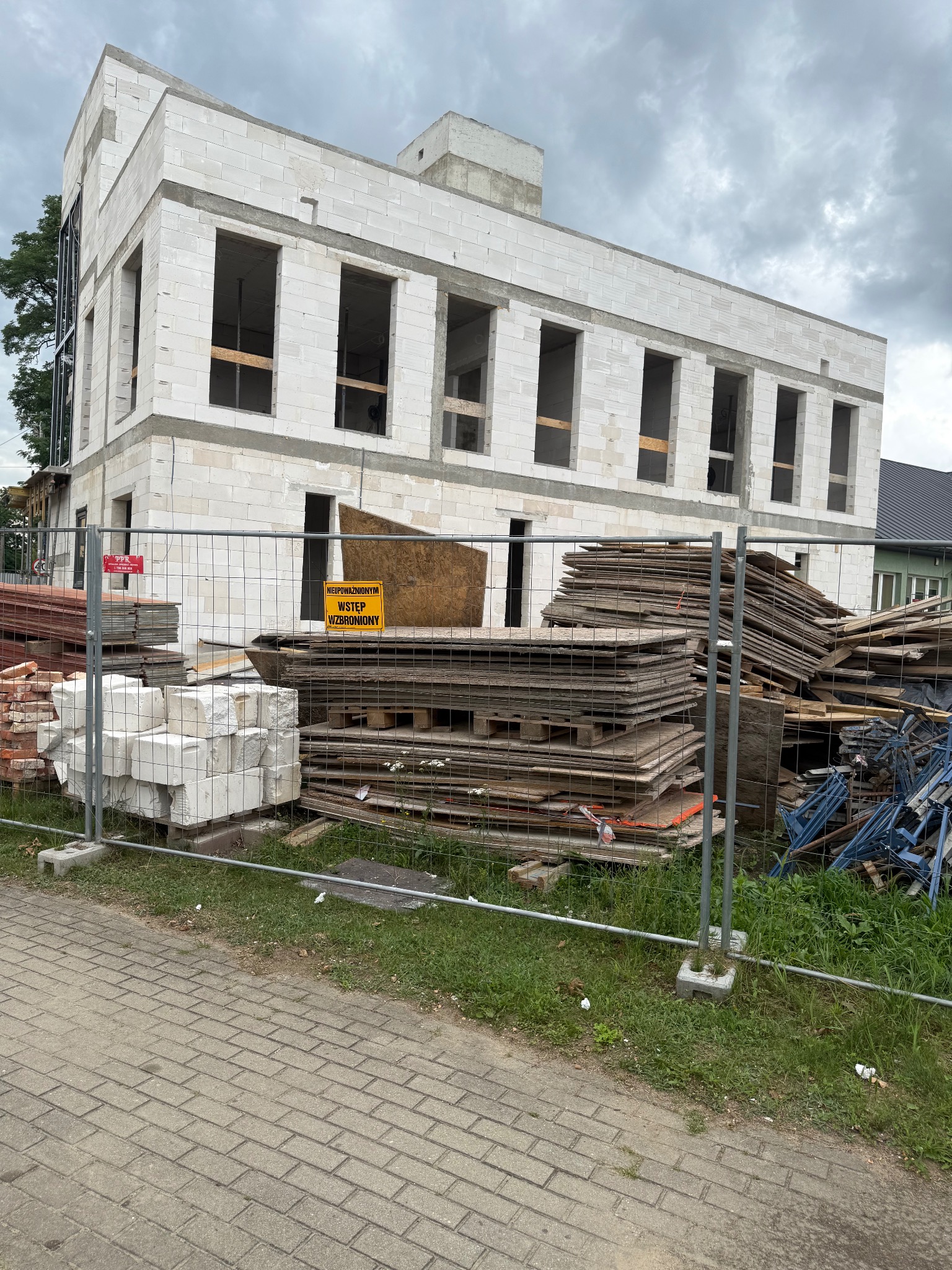 Dwukondygnacyjny budynek w stanie surowym, wzniesiony z białych bloczków, ogrodzony siatką, z widocznymi materiałami budowlanymi i tablicą ostrzegawczą.