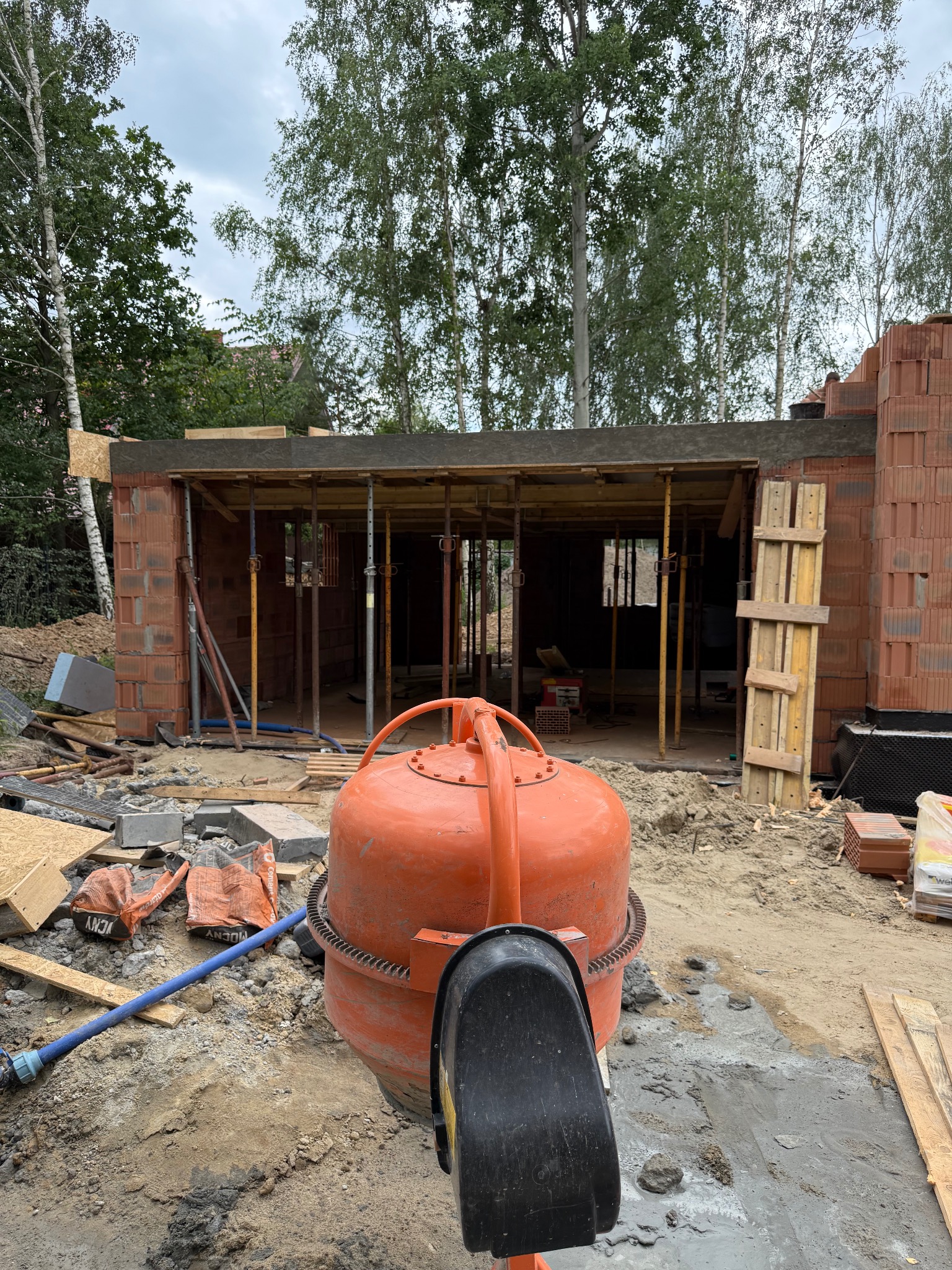 Budowa domu w trakcie: pomarańczowa betoniarka na pierwszym planie, w tle surowe ściany z czerwonej cegły i podpory stropu. Widoczne materiały budowlane i otoczenie placu budowy.