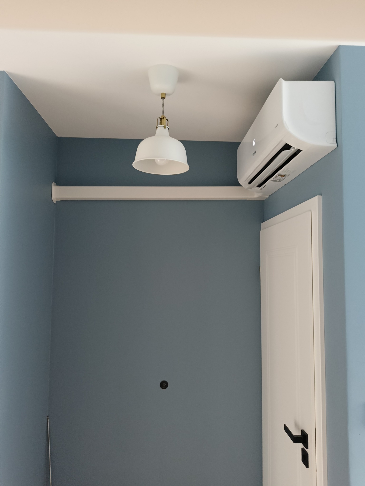 Biały klimatyzator Gorenje zamontowany nad drzwiami w pokoju o błękitnych ścianach, oświetlony lampą w stylu retro. Widoczna biała rura instalacyjna.