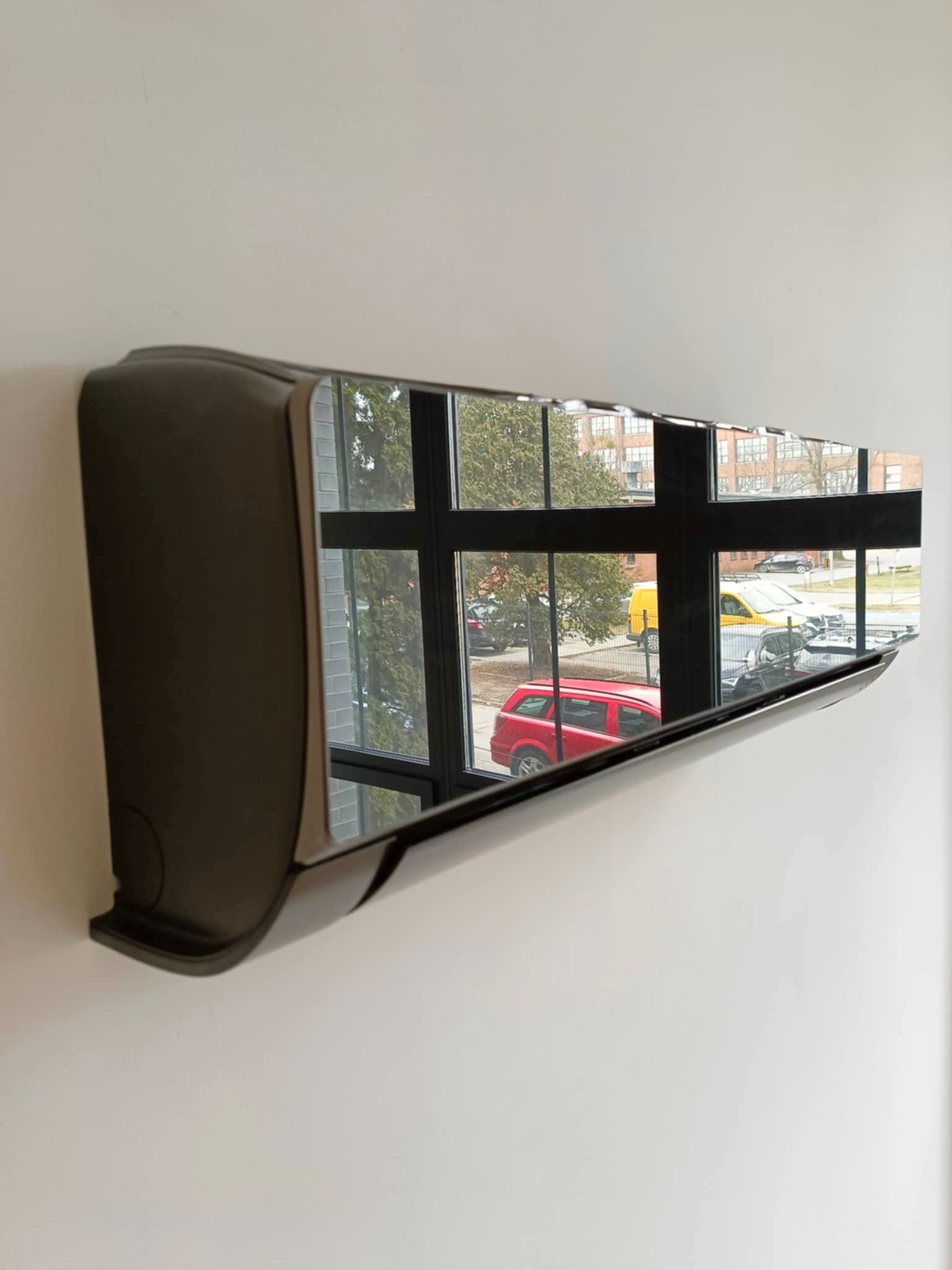 Czarny klimatyzator ścienny z widokiem na zewnątrz odbijającym się w błyszczącej obudowie, zamontowany na białej ścianie w nowoczesnym wnętrzu.