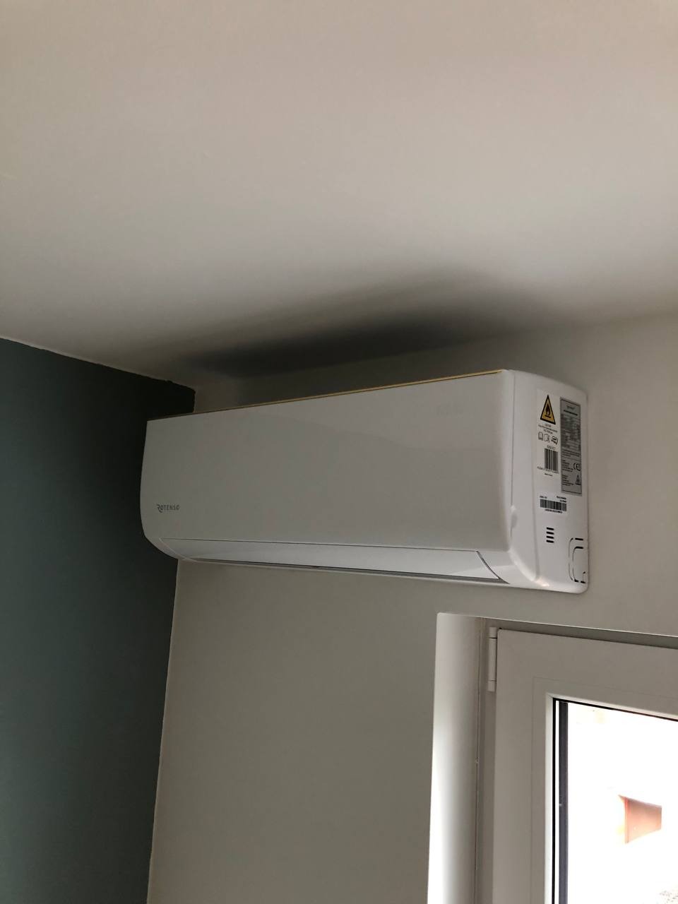 Biała klimatyzacja Rotenso zamontowana na ścianie nad oknem w jasnym pomieszczeniu. Widoczny fragment zielonej ściany po lewej stronie.