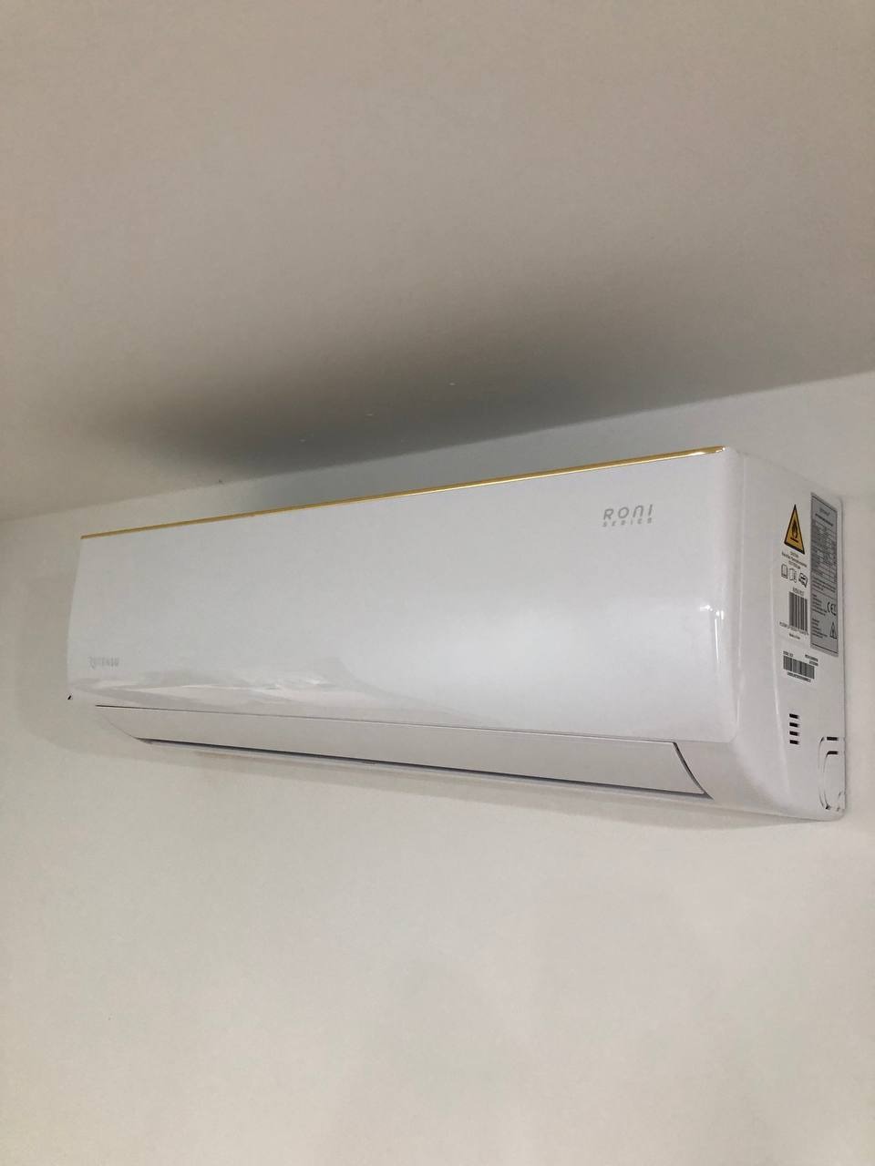 Biała klimatyzacja z subtelnym złotym akcentem na górnej krawędzi, zamontowana na jasnej ścianie w pomieszczeniu. Widoczne detale obudowy i oznaczenia producenta.