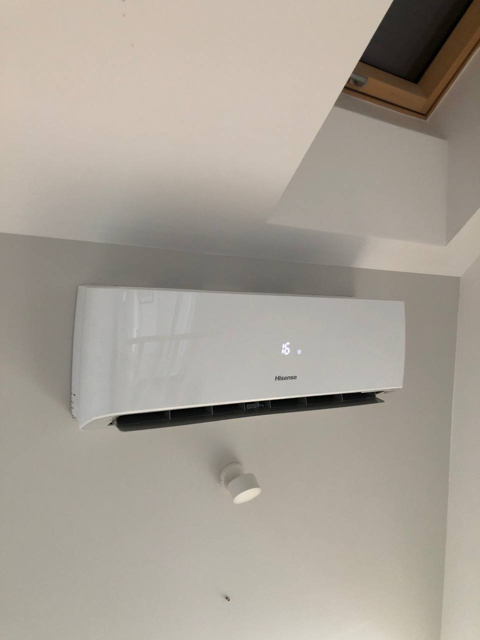 Biały klimatyzator Hisense zamontowany na szarej ścianie w pomieszczeniu z oknem dachowym. Widoczny wyświetlacz z temperaturą 16 stopni i oświetlenie punktowe.