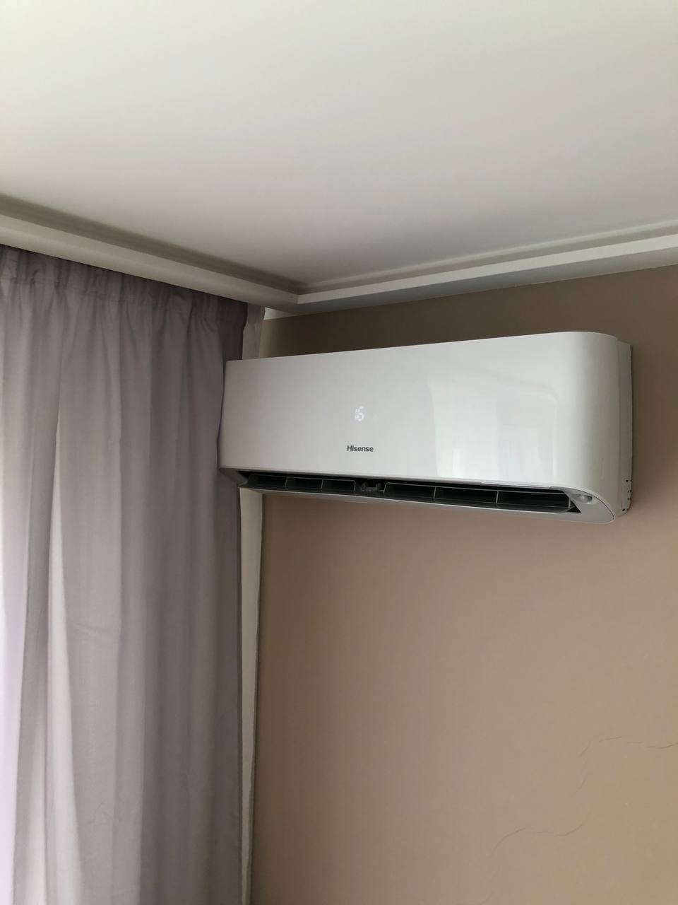 Biała klimatyzacja Hisense na beżowej ścianie w pokoju z zasłoniętym oknem. Widoczny fragment sufitu z ozdobną listwą. Minimalistyczny design.