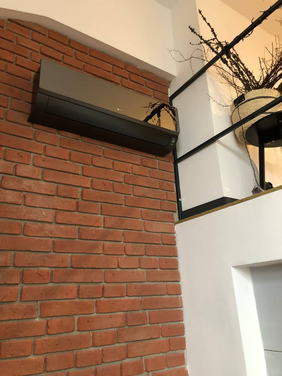 Ciemny klimatyzator LG zamontowany na ceglanej ścianie w nowoczesnym wnętrzu. Widoczny fragment dekoracji roślinnej i biała ściana.