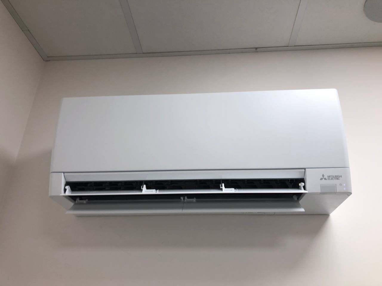 Biała, naścienna jednostka klimatyzacji Mitsubishi Electric, zamontowana w pomieszczeniu z podwieszanym sufitem kasetonowym. Widoczne otwarte żaluzje.