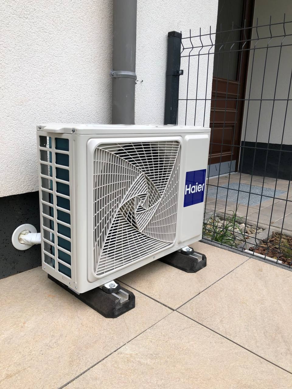 Zewnętrzna jednostka klimatyzacji Haier, zamontowana na betonowej podstawie przy budynku z rurą spustową. Widoczny fragment ogrodzenia panelowego.