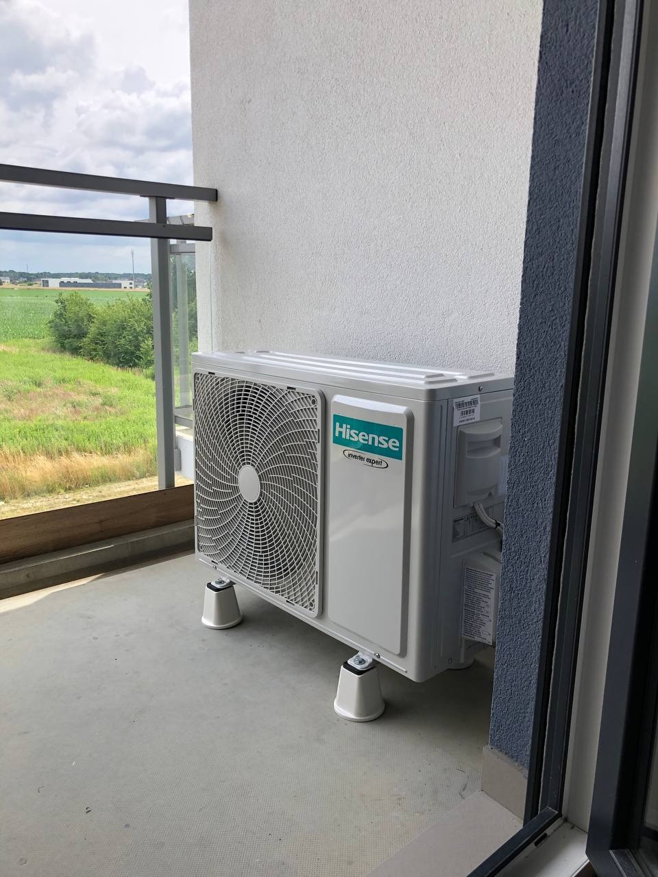 Biała, zewnętrzna jednostka klimatyzacji Hisense na balkonie z widokiem na zielone pola. Urządzenie stoi na białych, podwyższających nóżkach.