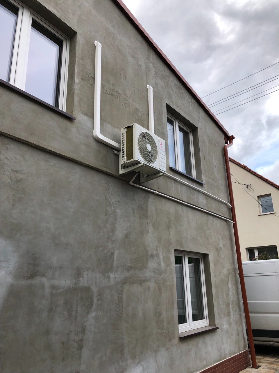 Zewnętrzny klimatyzator zamontowany na szarej ścianie budynku z widocznymi białymi rurami odprowadzającymi. Ujęcie z dołu, pochmurne niebo w tle.