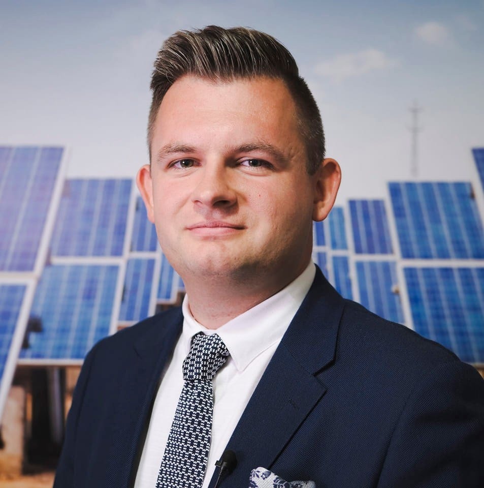 Mężczyzna w garniturze na tle paneli słonecznych, z mikrofonem krawatowym. Uśmiechnięty, profesjonalny wizerunek w kontekście energii odnawialnej.