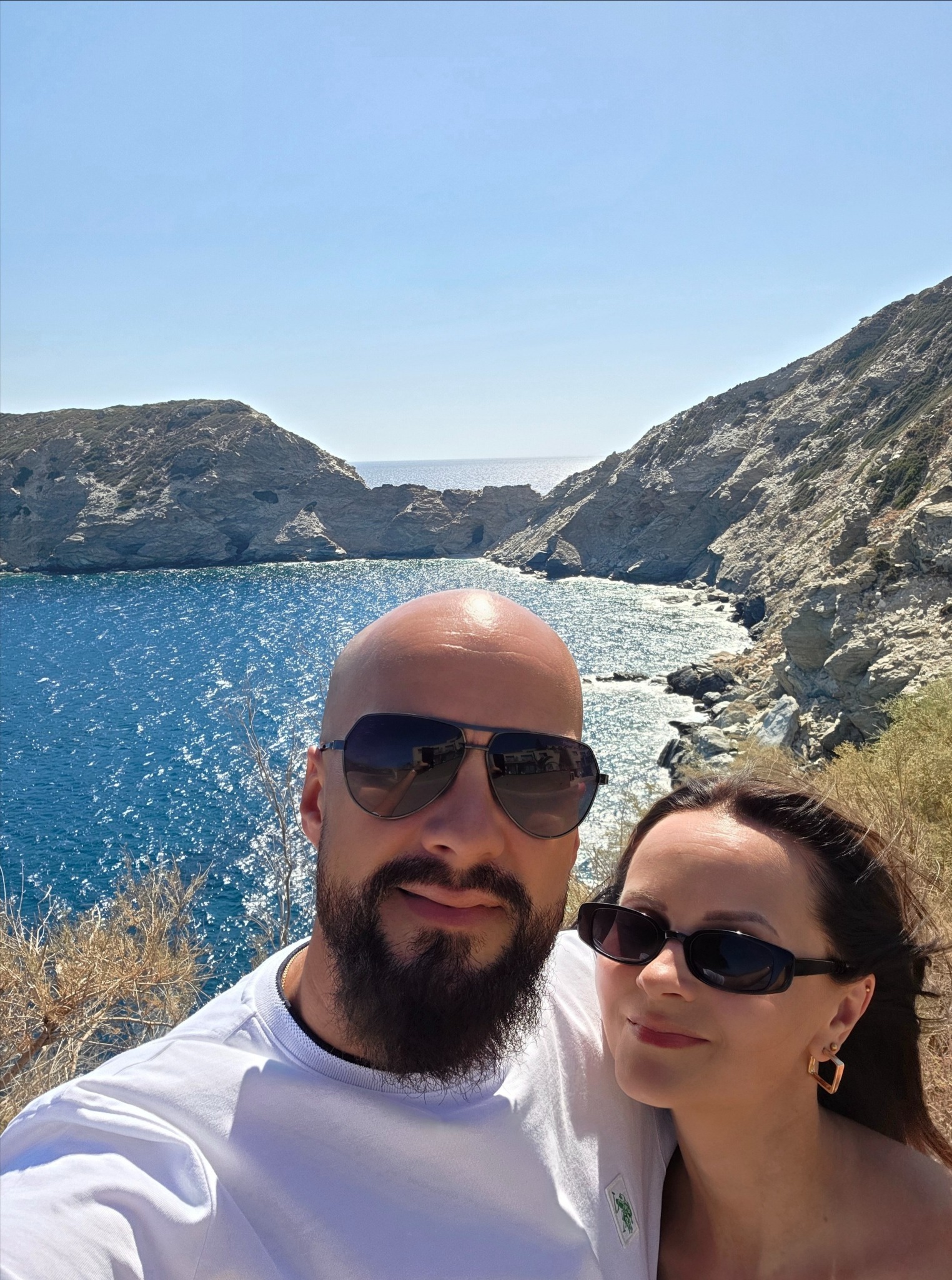 Uśmiechnięta para w okularach przeciwsłonecznych pozuje na tle malowniczej zatoki z klifami i błękitnym morzem. Jasne, słoneczne oświetlenie.