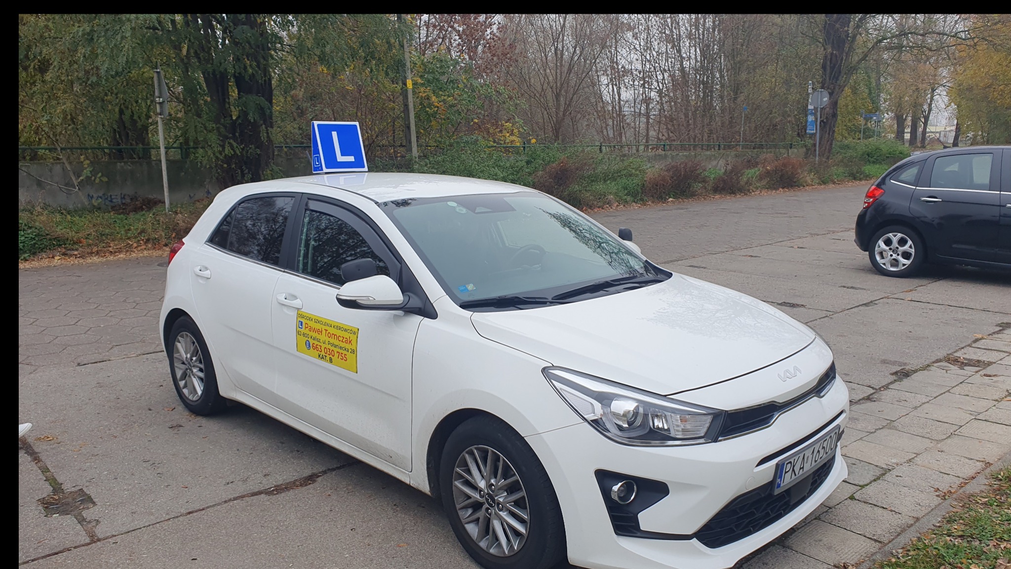 Biała Kia Rio z oznaczeniem 'L' na dachu, zaparkowana na betonowym placu. Na drzwiach naklejka z danymi szkoły jazdy 'Paweł Tomczak'.