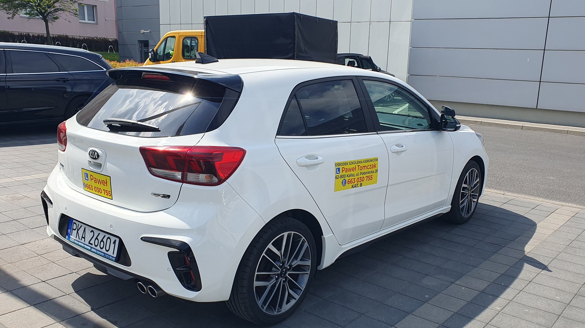 Biała Kia Rio z naklejkami szkoły jazdy 'Paweł Tomczak' na karoserii, zaparkowana na chodniku przed budynkiem. Widoczny numer telefonu i adres.