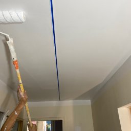 Nurii S.C. - Malowanie sufitu wałkiem na długim uchwycie; widoczne ślady farby na rękach malarza i niebieska taśma malarska oddzielająca pomalowaną i niepomalowaną część sufitu.
