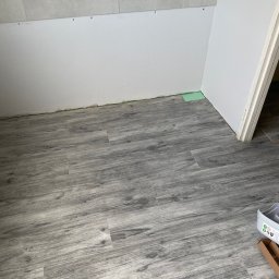 Nurii S.C. - Szare panele podłogowe imitujące drewno, ułożone w rogu pomieszczenia. Częściowo widoczna ściana z płyt kartonowo-gipsowych i fragment listwy przypodłogowej.
