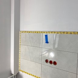 Nurii S.C. - Świeżo położone, jasne płytki w łazience, zabezpieczone taśmą malarską, z widocznymi otworami pod instalację elektryczną. Minimalistyczny design, w trakcie wykańczania.