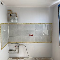 Nurii S.C. - Fragment łazienki w trakcie prac: częściowo ułożone szare płytki, zabezpieczone taśmą, instalacja gazowa i hydrauliczna. Podłoga z resztkami zaprawy.