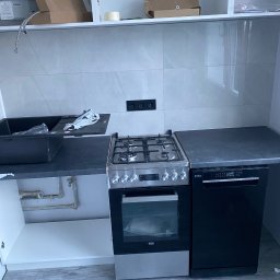 Nurii S.C. - Nowoczesna kuchnia w trakcie montażu: czarny zlew, płyta gazowa, piekarnik Beko i zmywarka Amica. Szare płytki na ścianie, drewnopodobna podłoga.