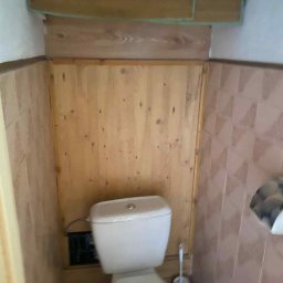 Nurii S.C. - Kompakt WC w obudowie z boazerii w małej łazience z brązowymi płytkami. Widoczna toaleta, ściany wyłożone drewnem i płytkami oraz szafka wisząca.