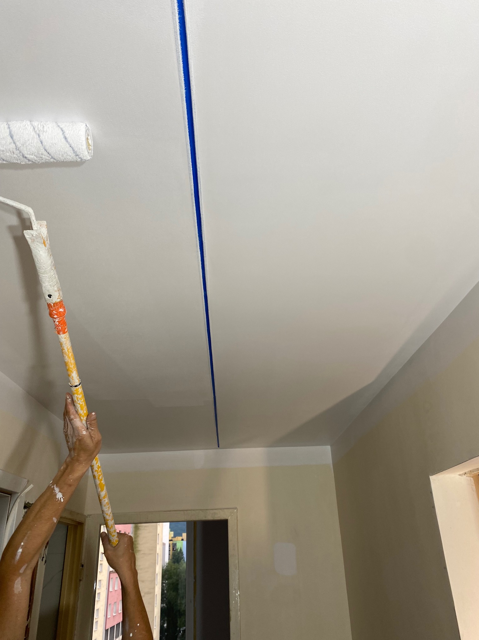Malowanie sufitu wałkiem na długim uchwycie; widoczne ślady farby na rękach malarza i niebieska taśma malarska oddzielająca pomalowaną i niepomalowaną część sufitu.