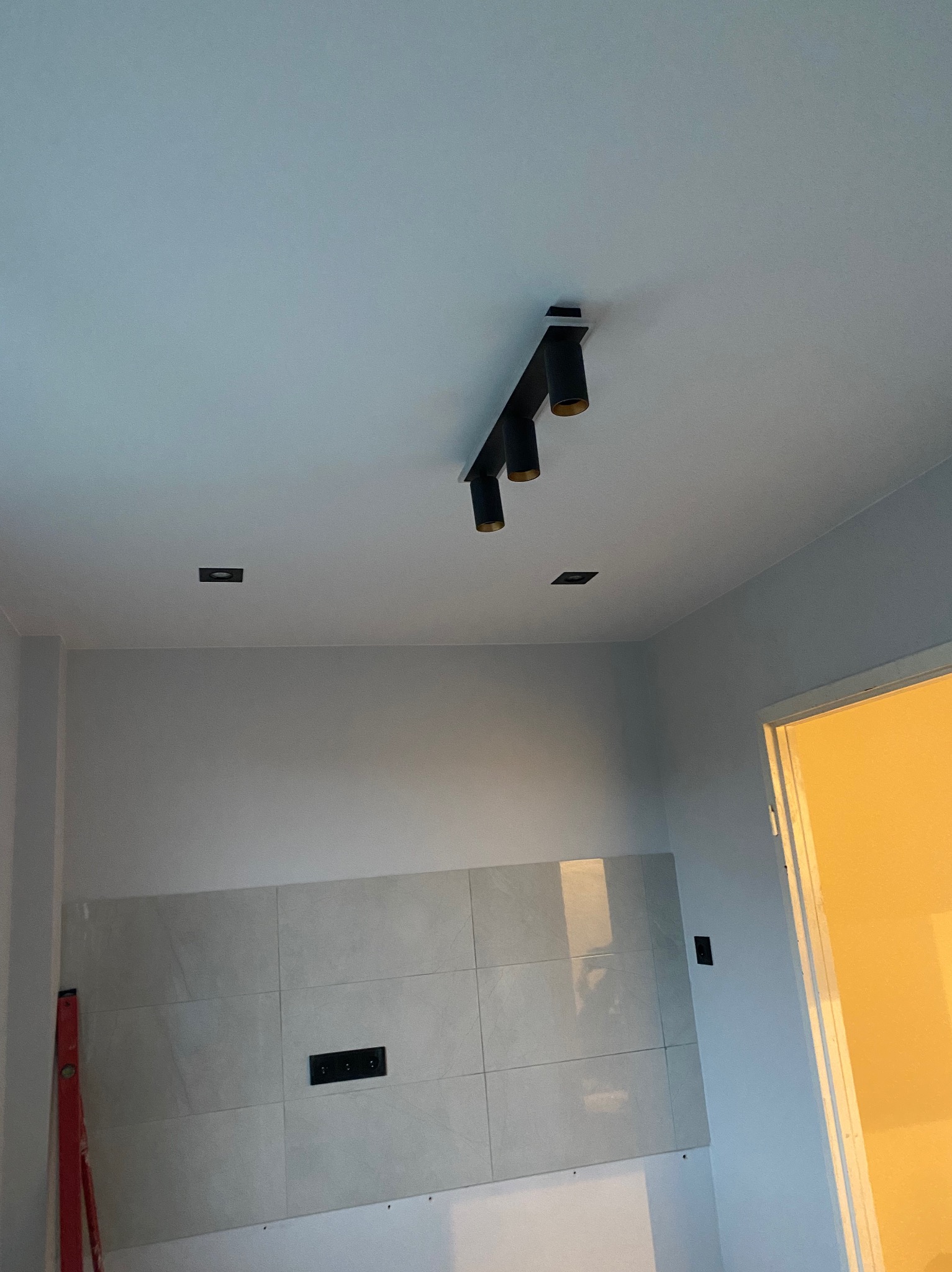 Ściana w trakcie wykańczania z szarymi płytkami, widoczne czarne punkty świetlne i poziomica przy ścianie, fragment otwartych drzwi z żółtym światłem.
