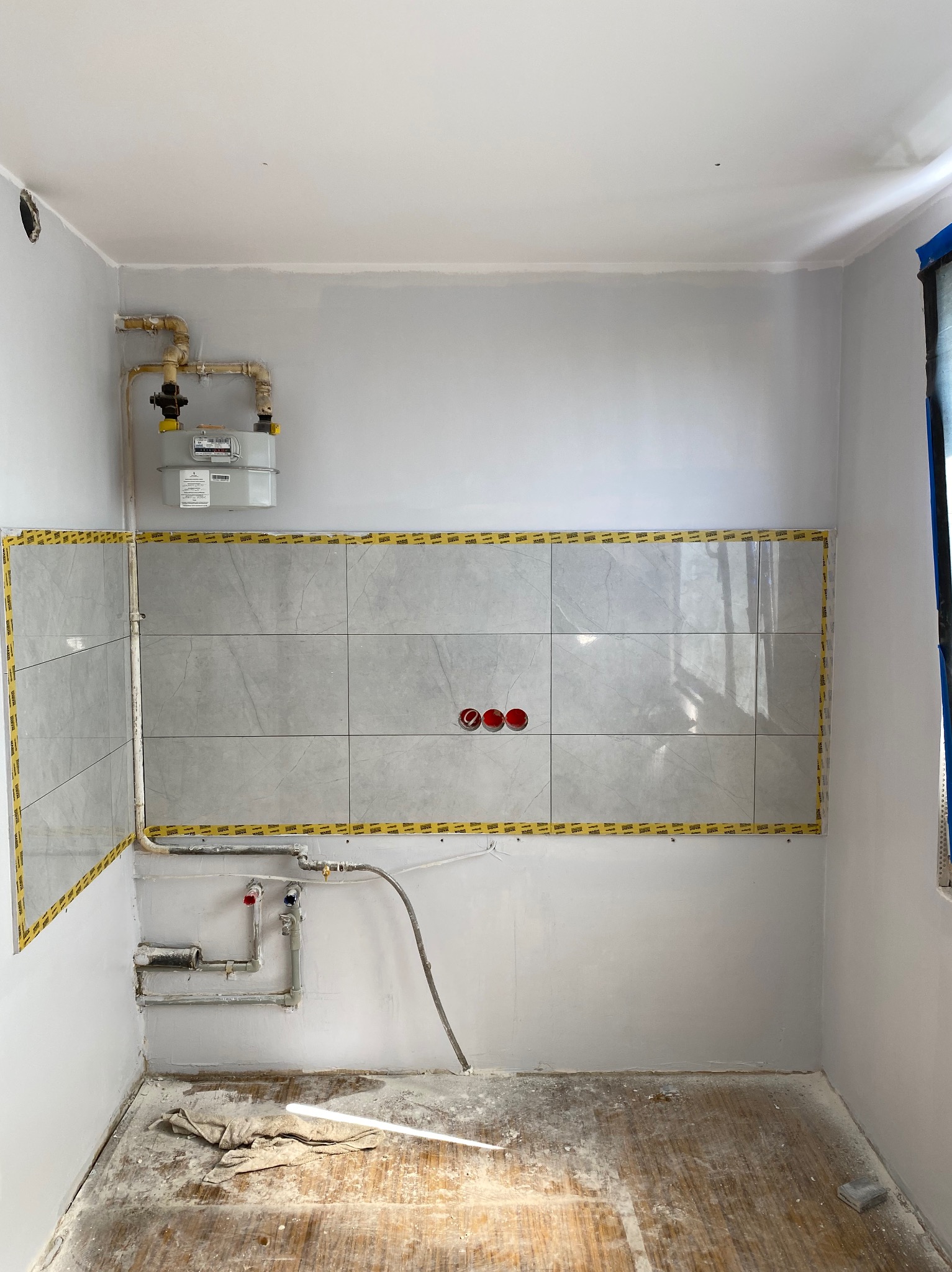Fragment łazienki w trakcie prac: częściowo ułożone szare płytki, zabezpieczone taśmą, instalacja gazowa i hydrauliczna. Podłoga z resztkami zaprawy.