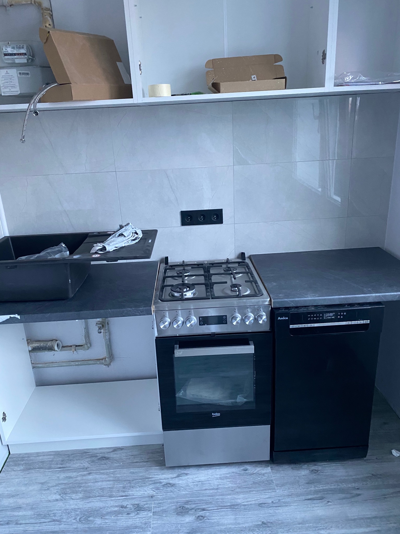 Nowoczesna kuchnia w trakcie montażu: czarny zlew, płyta gazowa, piekarnik Beko i zmywarka Amica. Szare płytki na ścianie, drewnopodobna podłoga.