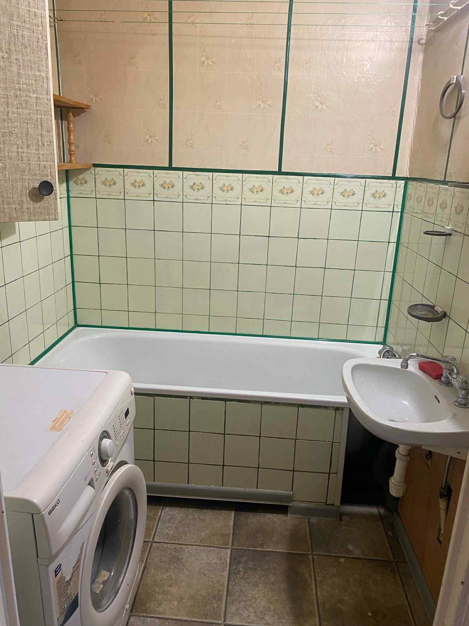 Łazienka z wanną, umywalką i pralką w bloku. Kremowe płytki z zielonymi akcentami. Widok z perspektywy podłogi, ukazujący stan pomieszczenia.