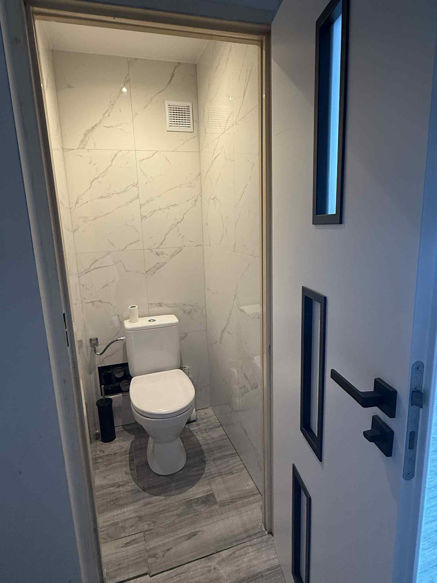 Wnętrze wykończonej łazienki z toaletą, wyłożonej marmurowymi płytkami. Widoczna biała toaleta, szare płytki podłogowe imitujące drewno i białe drzwi z czarnymi akcentami.